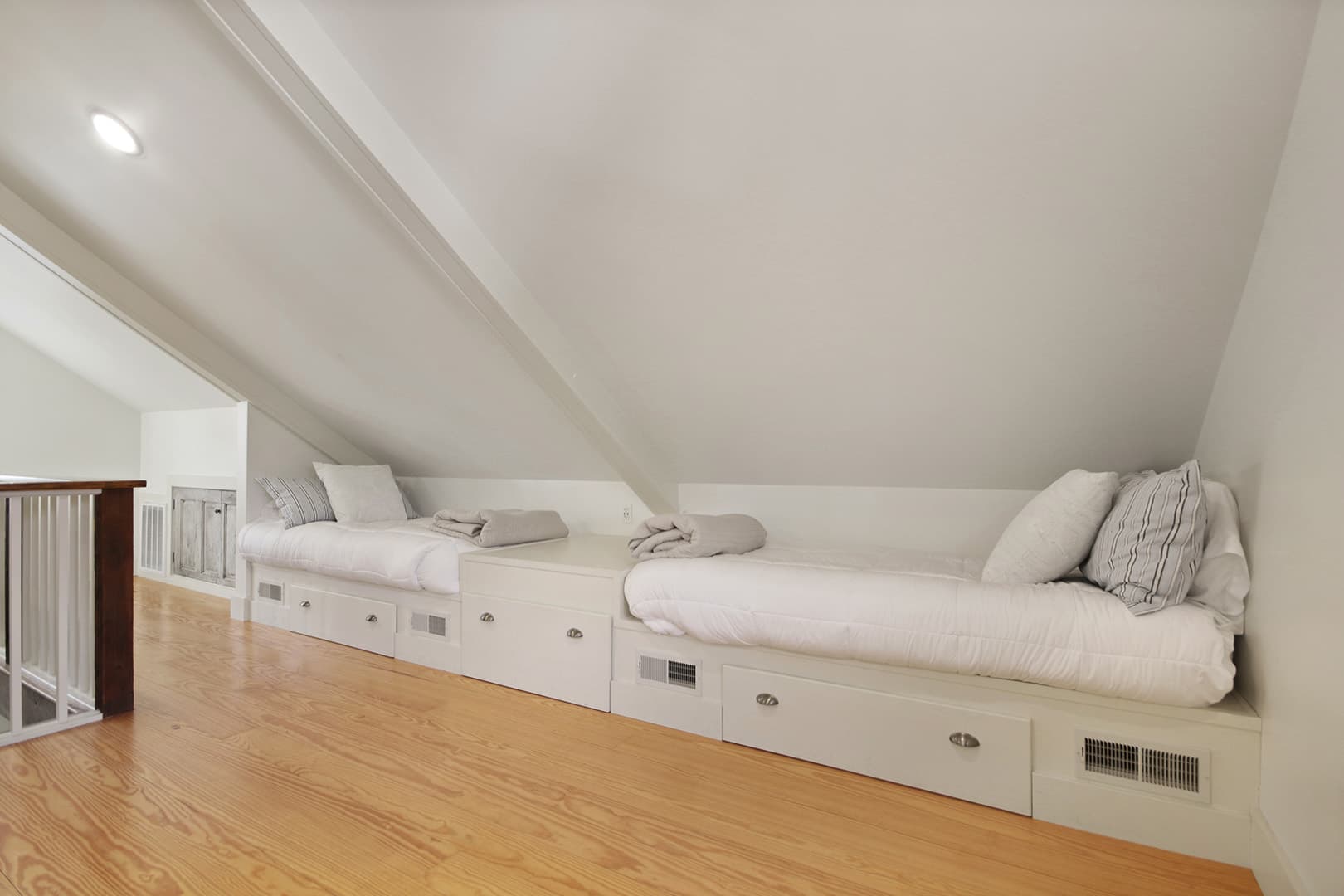 A charming attic bedroom featuring two built-in beds with white bedding, clever storage drawers, and a light wood floor under a sloped ceiling.