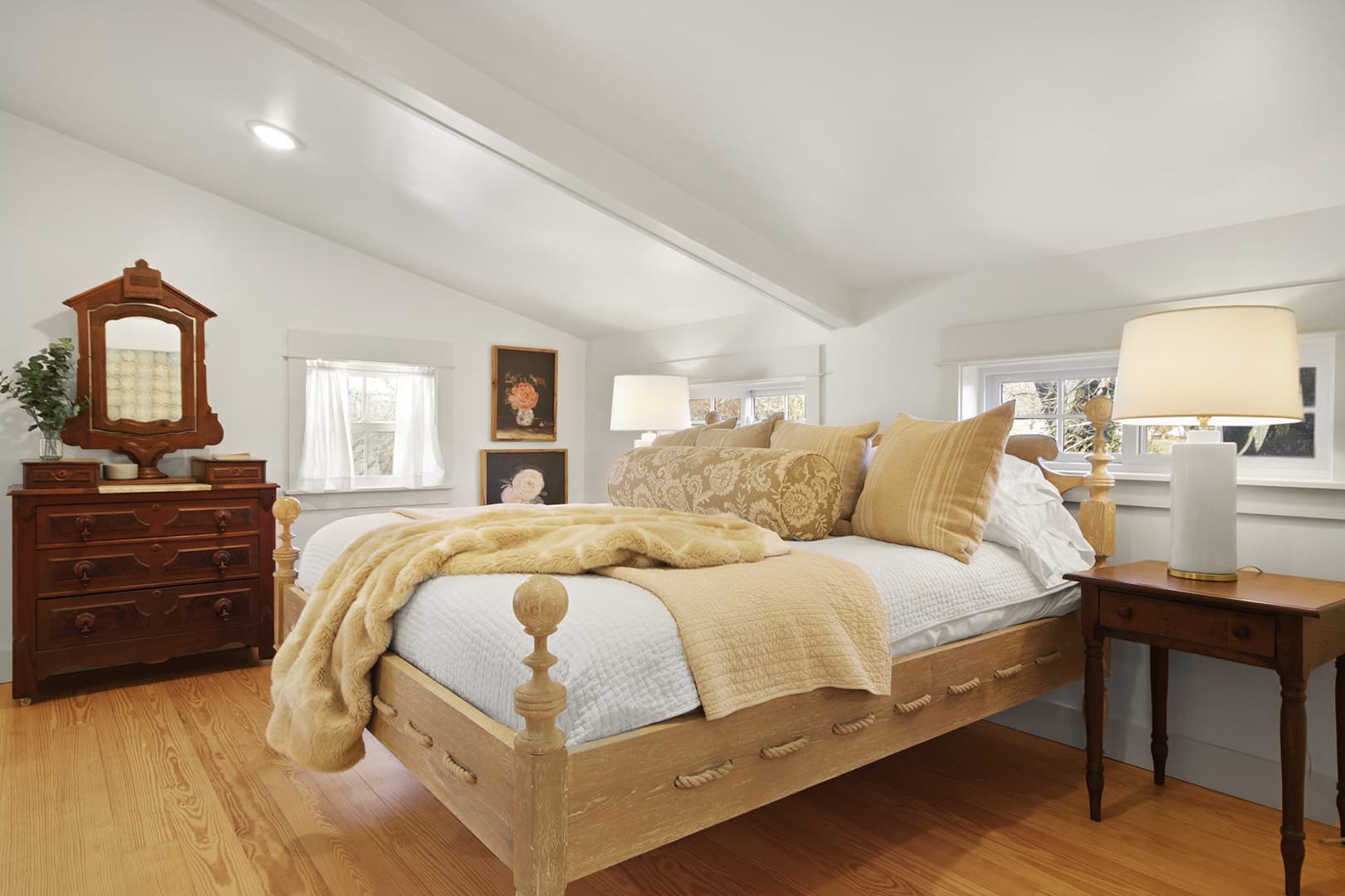 A bright, cozy attic bedroom with a light wood bed, warm-toned bedding, a vintage dresser, and two bedside lamps.