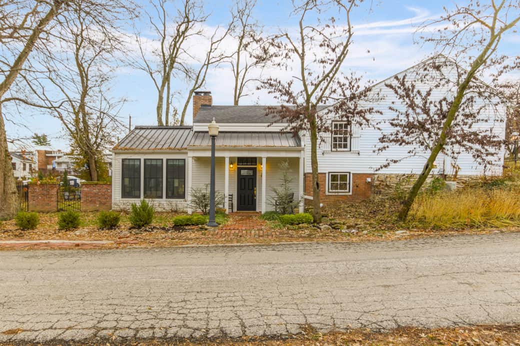 A charming, traditional house with a white facade and a covered porch, set amidst trees with bare branches under a partly cloudy sky.
