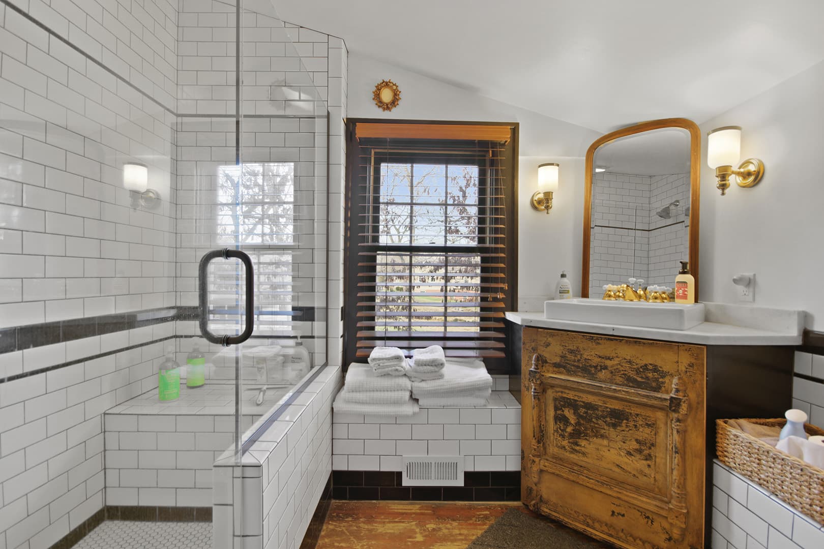 A bright bathroom featuring a white subway-tiled shower, a unique wooden vanity, and a large window with blinds.