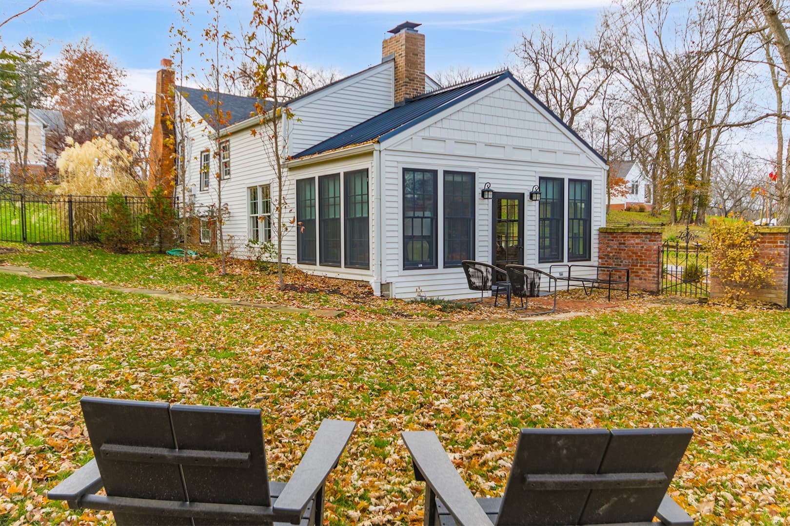 A charming white house with a dark roof and a sunroom addition, nestled in a leafy yard with two Adirondack chairs facing the home in the foreground.