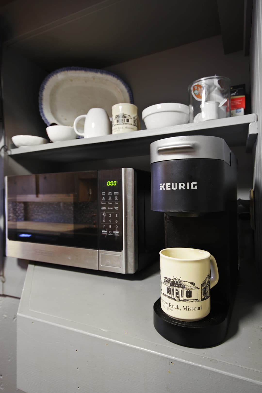 A microwave, a Keurig coffee maker, and white dishes on open shelving.