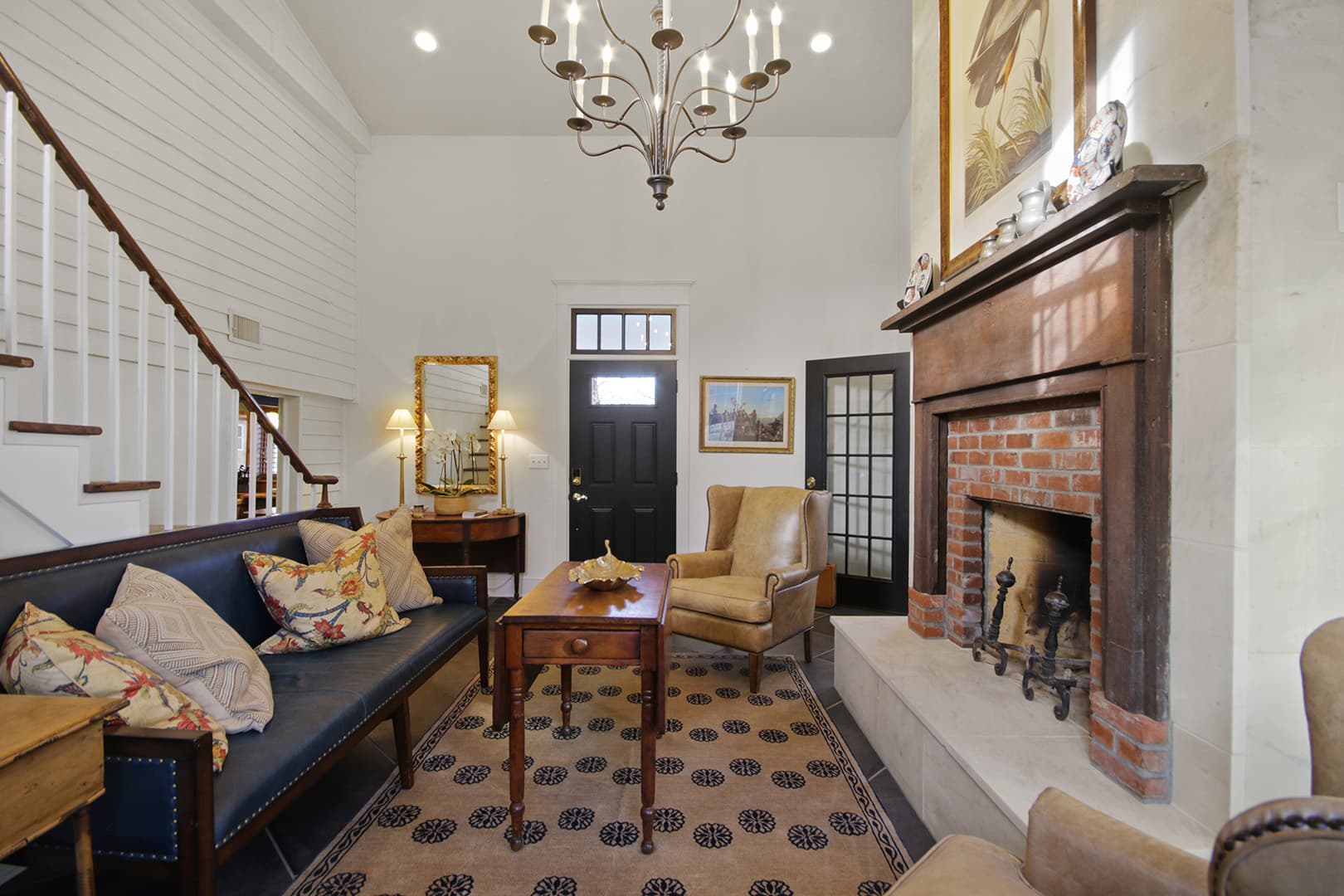 A classic living room with a fireplace, dark wood mantel, high ceilings, a grand chandelier, and a prominent staircase.