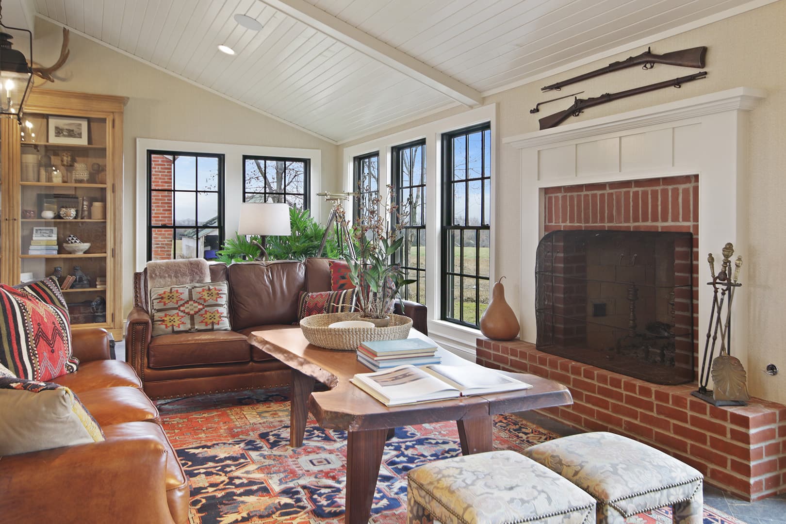 An inviting great room with a rustic farmhouse aesthetic, featuring a brick fireplace, leather sofas, a vaulted ceiling, and ample windows.