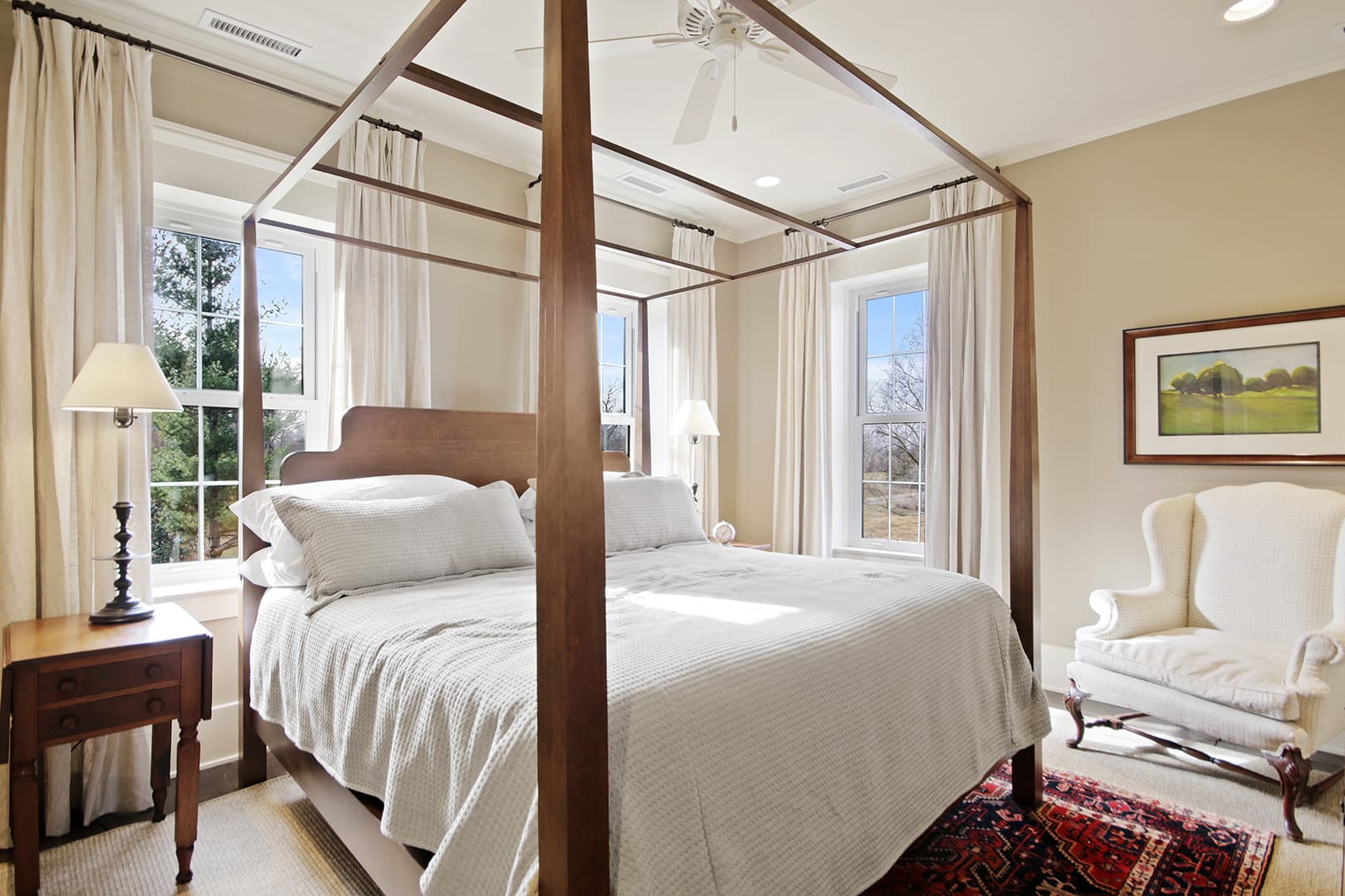 A serene bedroom with a dark wood four-poster bed, white bedding, an oriental rug, and a comfortable armchair.