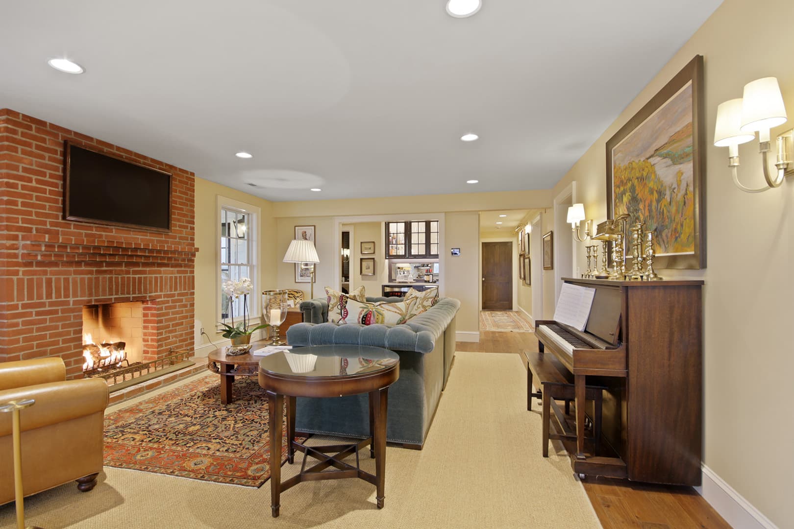 A warm, traditional living room with a brick fireplace, comfortable seating, a piano, and elegant lighting.