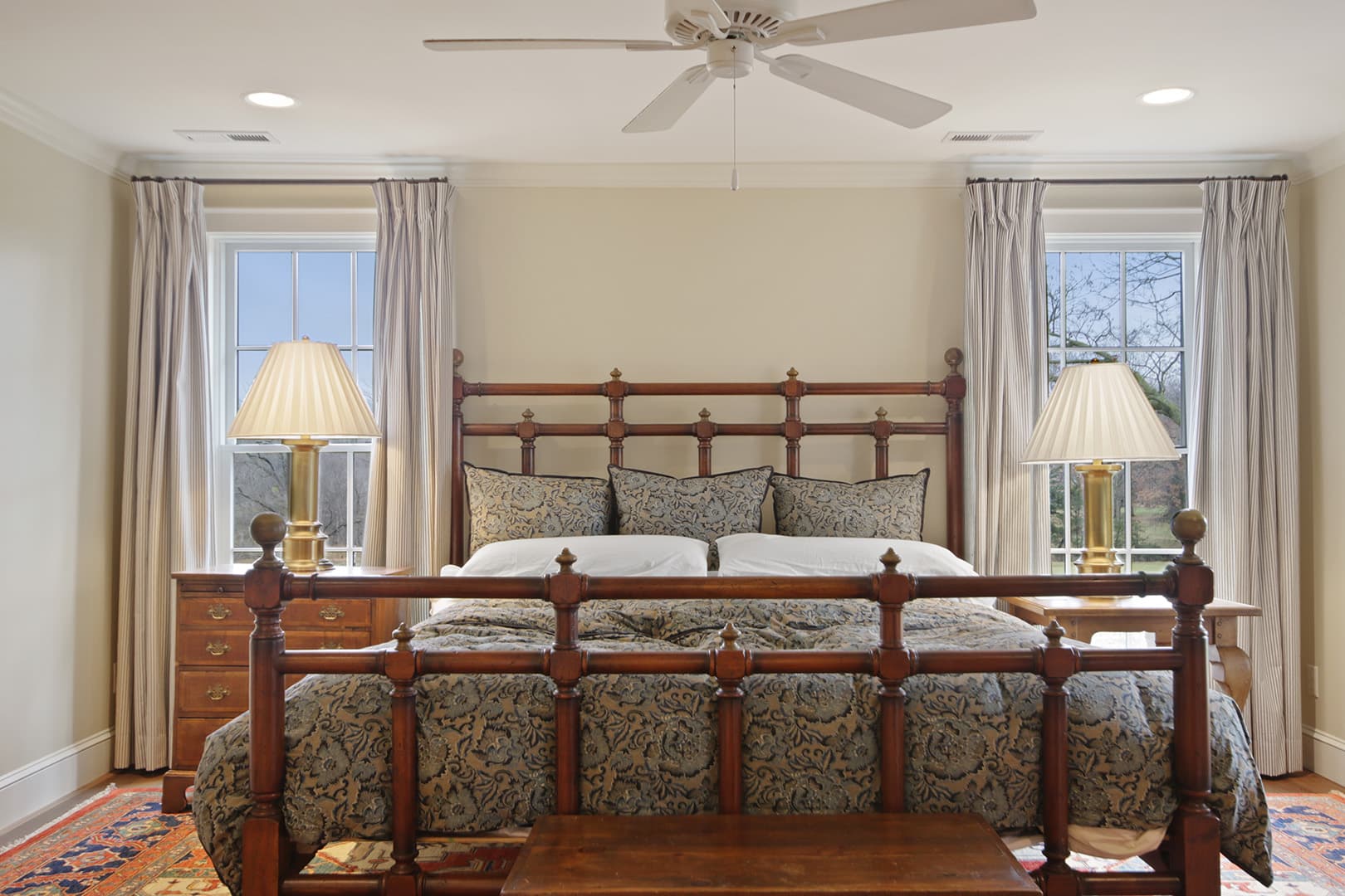 A serene bedroom featuring a traditional wooden bed frame, patterned bedding, two matching bedside lamps, and a ceiling fan.